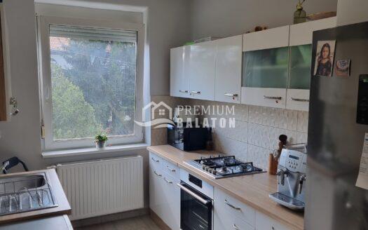Modern kitchen with white glossy upper and lower cabinets, a light wood countertop, and stainless steel appliances including a built-in oven and gas cooktop; a window over the sink brings in natural light, with a small plant on the windowsill and a round table edge in the foreground.