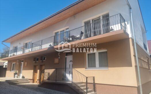 Exterior view of a multi-story beige building with decorative black wrought-iron balconies and steps leading to a front entrance against a clear blue sky. Watermark reads 'Premium Balaton' across the lower center.