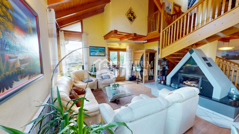 Bright living room with cream sofas, glass coffee table, and indoor plants under a wooden vaulted ceiling and stairs.