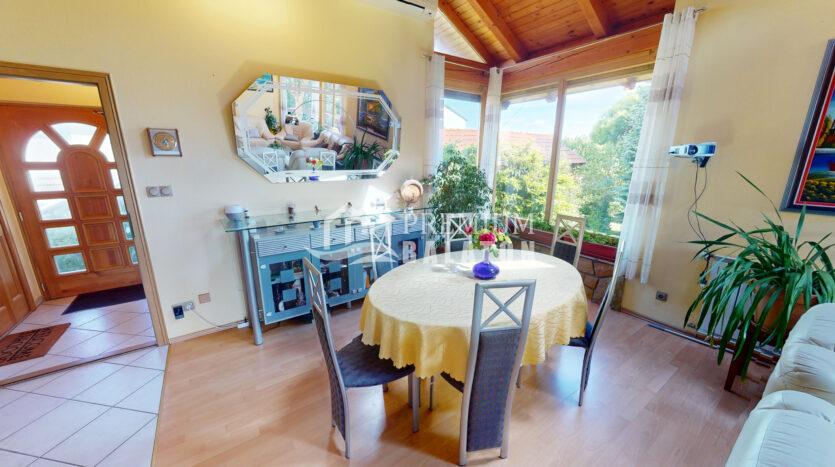 Bright dining area with a round table covered by a yellow tablecloth, a blue sideboard, and large windows overlooking greenery.