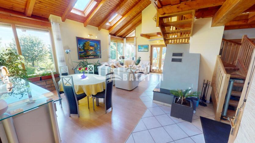 Open-plan living and dining area with exposed wooden beams, large windows, and a staircase to the right; plants and tropical artwork add color.