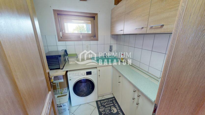 Small laundry room with a front-loading washing machine under a light countertop and wooden upper cabinets on the right, tiled backsplash, and a window with a wooden frame on the left.