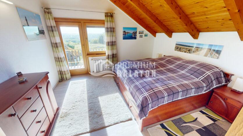 Cozy bedroom with a plaid bedspread, wooden furniture, and a sloped wooden ceiling; large window and balcony door overlook a green landscape.