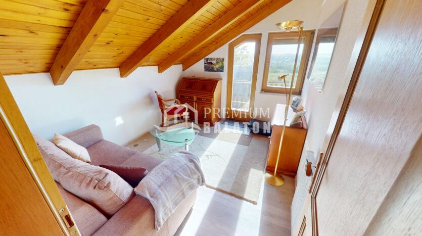 Cozy attic living room with a beige sofa, glass coffee table, wooden beams, and a large window overlooking a hillside valley.