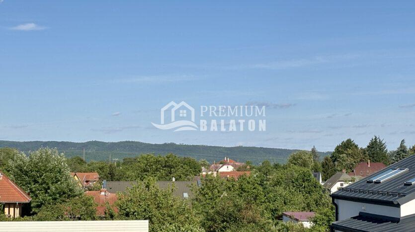 Suburban neighborhood with red-tiled roofs, green trees, and distant hills under a clear blue sky.
