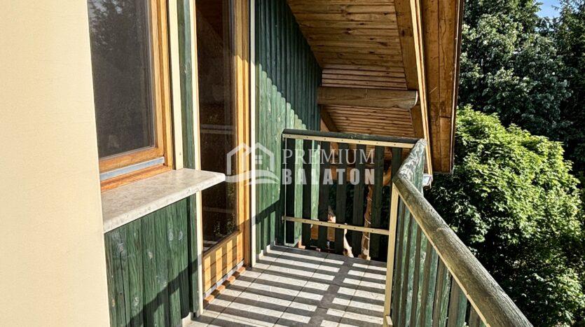 Balcony along a green wooden wall with beige trim, tiled floor, and a wooden railing, overlooking trees and a blue sky.