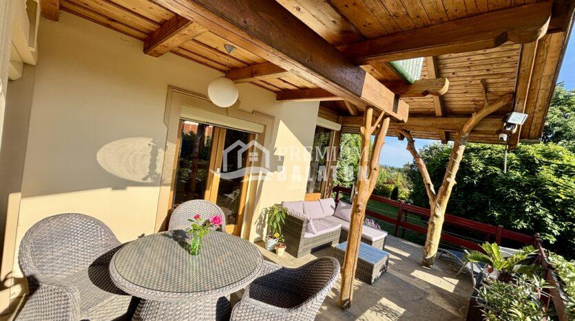 Outdoor patio seating area with round wicker table and matching chairs under a timber-beamed pergola, sunshine, and potted plants nearby.