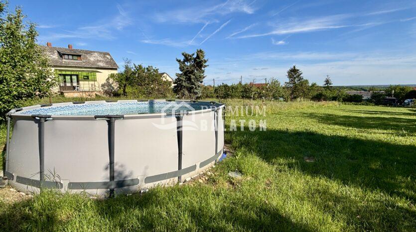 Above-ground round pool in a grassy backyard, with a beige house on the left and a blue sky with contrails.