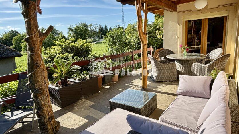 Sunny outdoor patio with wicker sectional sofa, cream cushions, and a glass coffee table on a stone floor; a wooden pergola and potted plants surround the seating area with a green garden beyond and a blue sky above