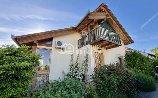 Two-story house with exposed wooden beams and a balcony, surrounded by lush green shrubs under a clear blue sky.
