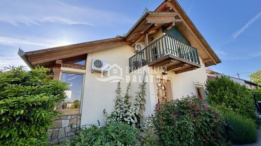 Two-story house with exposed wooden beams and a balcony, surrounded by lush green shrubs under a clear blue sky.