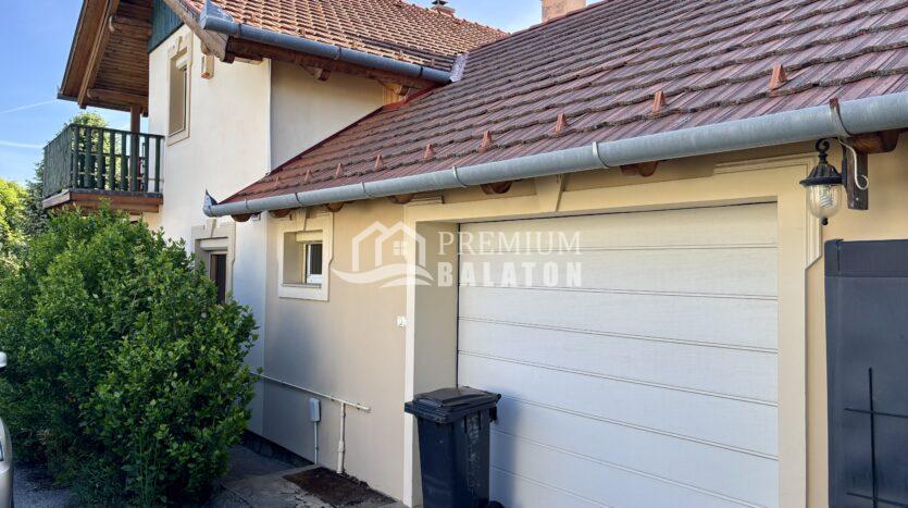 Exterior view of a beige house with a red-tiled roof, gutter, and a driveway with two black trash bins nearby.