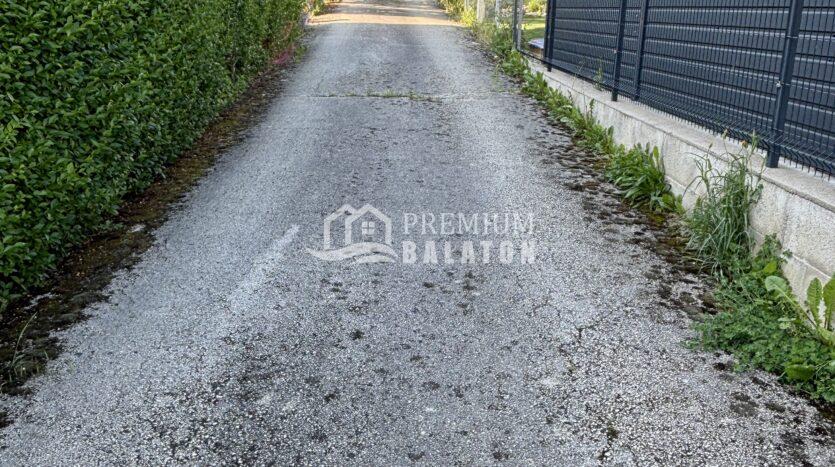 Narrow gravel alley flanked by a hedge on the left and a fence on the right, with string lights overhead and a car parked at the far end.