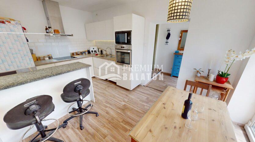 Bright open-plan kitchen with white cabinets, built-in oven and microwave, stainless sink, and a wooden dining table with wine and two glasses nearby.