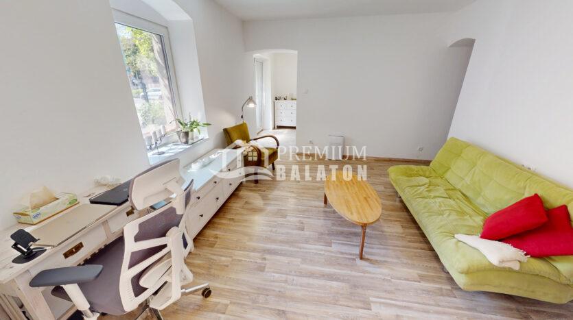 Bright home office and living area with a white desk, ergonomic chair, and a green sofa with red cushions nearby window light.