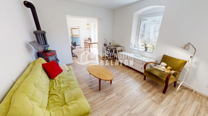 Bright living space with a lime-green sofa, red cushion, and wooden coffee table; a tall stove/chimney on the left. In the background is a desk area and a doorway to another room with a blue dresser.