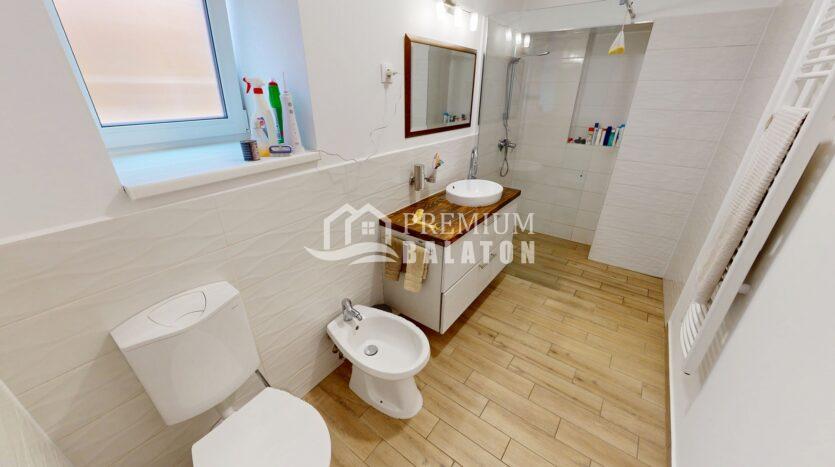 Bright white bathroom with a wooden countertop sink, mirror, and glass shower enclosure; window on the left and towel rack on the right.