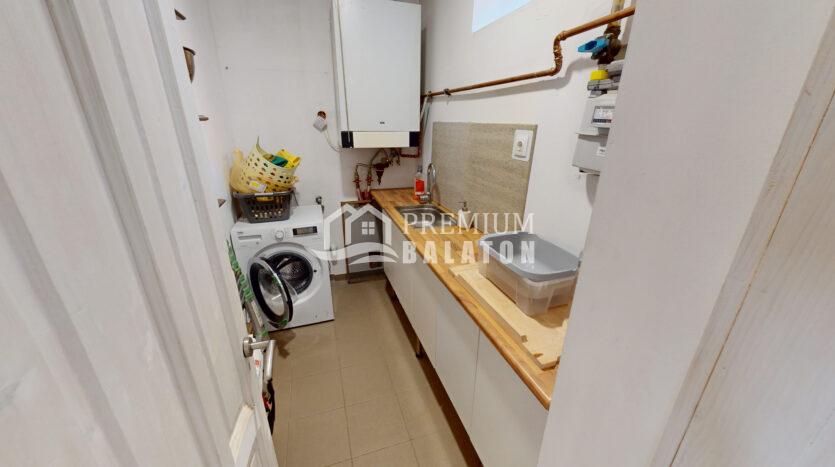 Narrow laundry room with a front-loading washing machine and a laundry basket on top, beside a wooden countertop and sink along white walls.