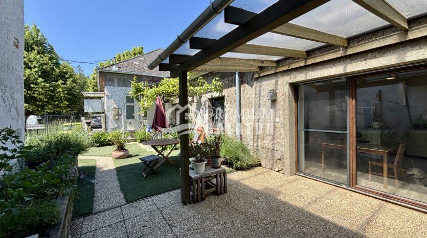 Backyard patio with a wooden pergola, clear roof panels, and sliding glass doors, surrounded by potted plants and greenery.
