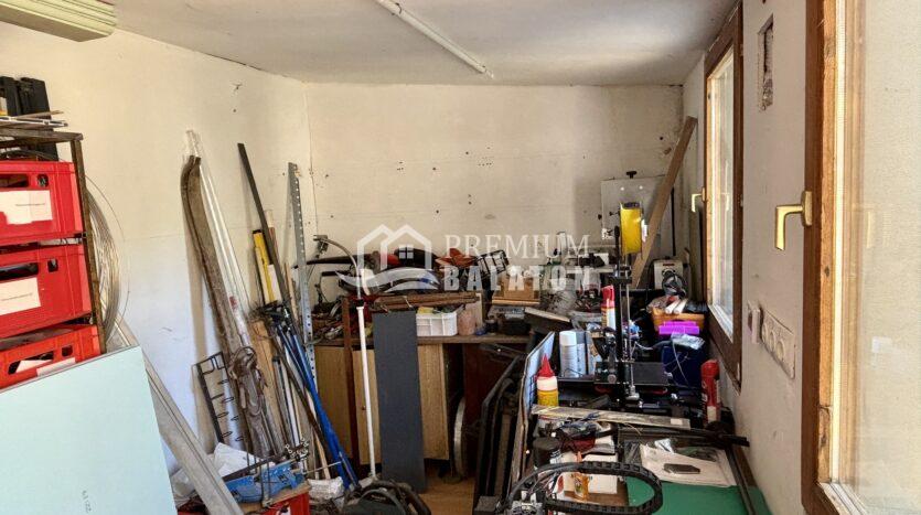 Cluttered workshop or garage with tools, metal shelving, cables, and random equipment strewn across tables and floor by a windowed wall.