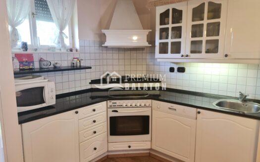 Small white kitchen with black countertops, built-in oven and microwave, and glass-front cabinets above a tiled backsplash under a white range hood. Window with sheer curtains to the left.